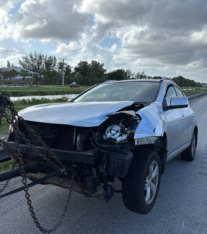 Silver SUV with heavy front-end accident damage attached to a tow truck chain, ready to sell a totaled car in Fort Lauderdale.