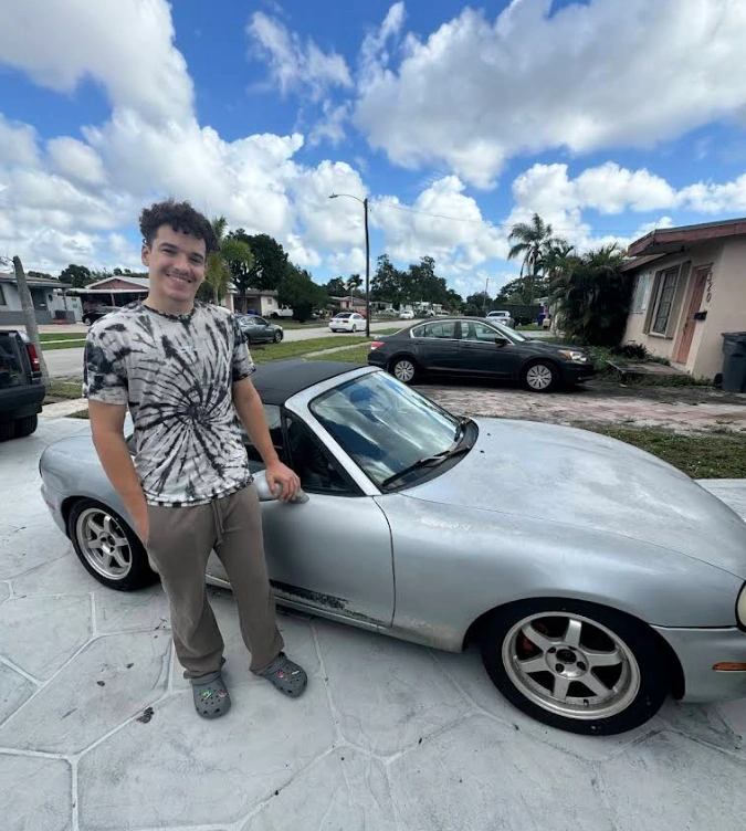 A young man smiling next to a silver convertible junk car with faded paint parked on a driveway, perfect for Junkyard Dogs's cash for junk cars in Miami services.