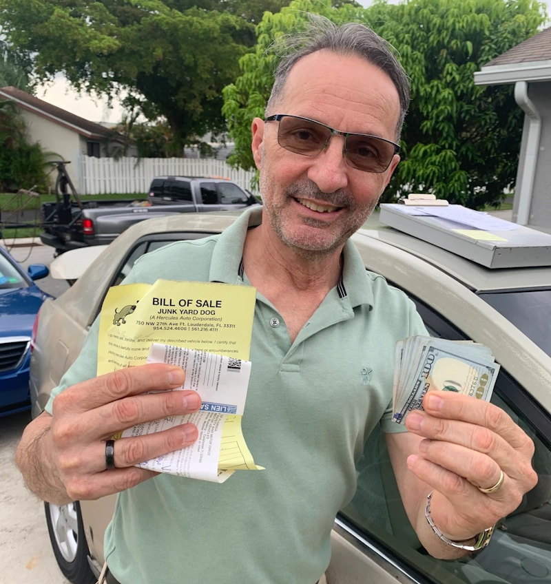 A man standing next to a car, holding a yellow bill of sale document and a stack of cash, illustrating key advice from junk car buyers in Boca Raton.
