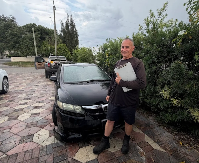 A man holding a clipboard stands next to a damaged black sedan, illustrating a service ready to buy broken car.