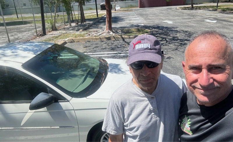 Two men standing in front of a white vehicle outdoors while discussing a car AC blowing hot air with Junkyard Dog.